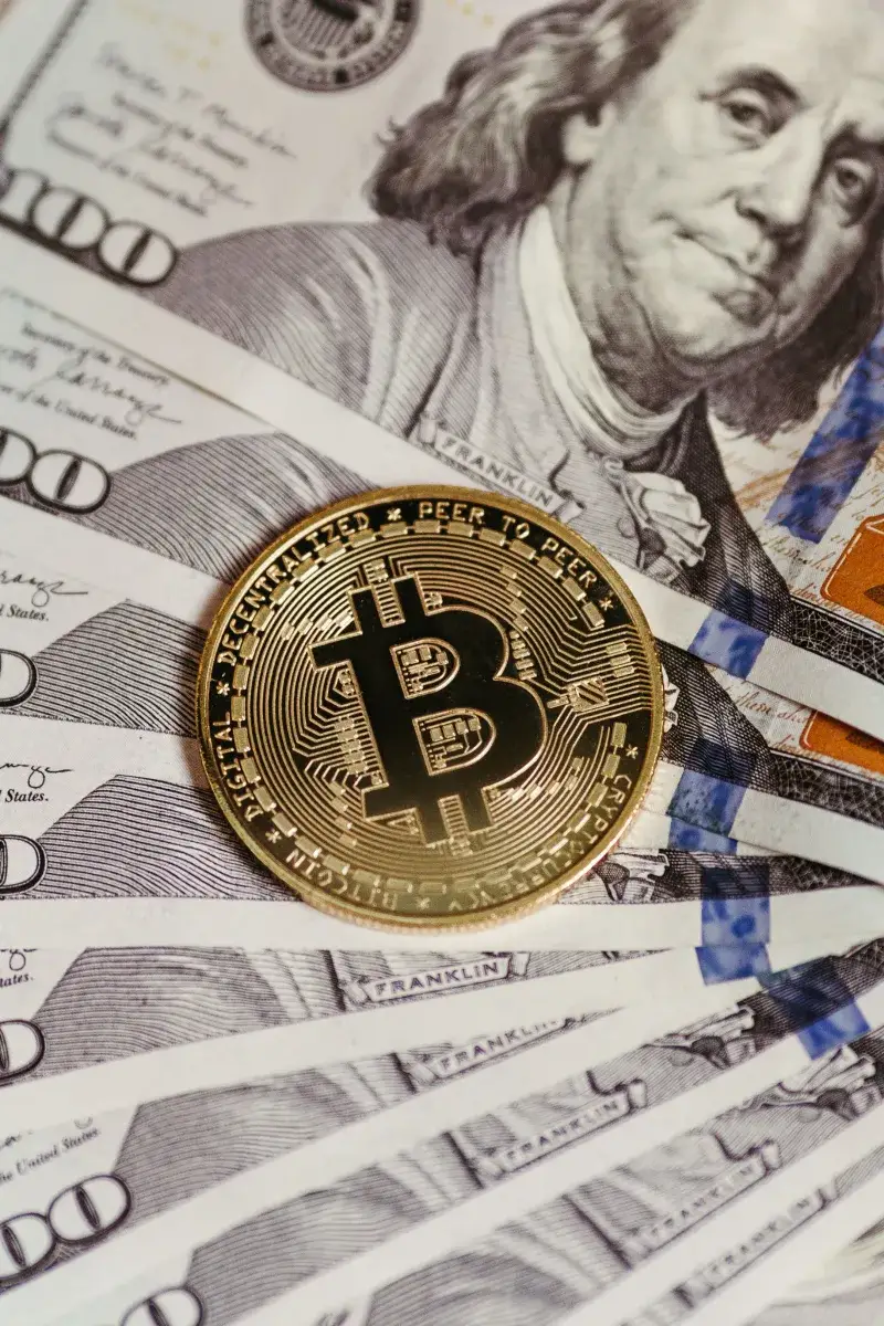 A vertical shot of a Bitcoin coin placed on multiple hundred-dollar bills symbolizing cryptocurrency and traditional finance.