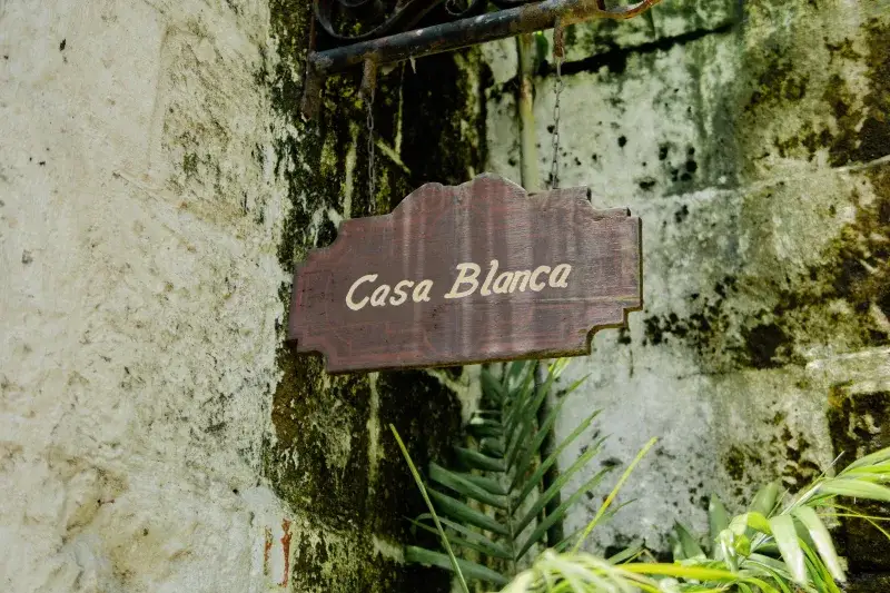 A wooden Casa Blanca sign hangs on a weathered stone wall surrounded by lush greenery.