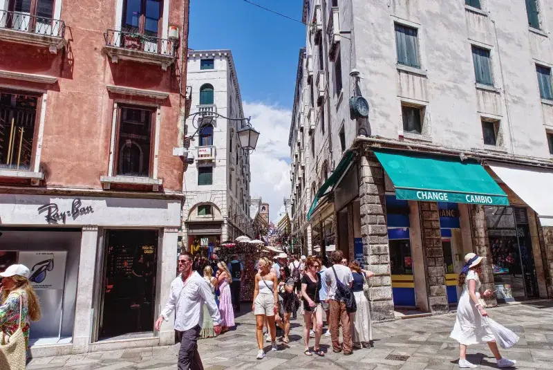 Lively street view in Venice with tourists exploring shops and architecture under a bright summer sky.