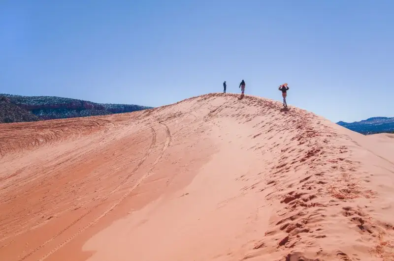 Three hikers atop the vibrant Coral Pink Sand Dunes in Utah under a clear blue sky.
