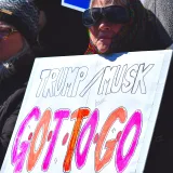 An older woman holds a protest sign against Trump and Musk outdoors in winter.