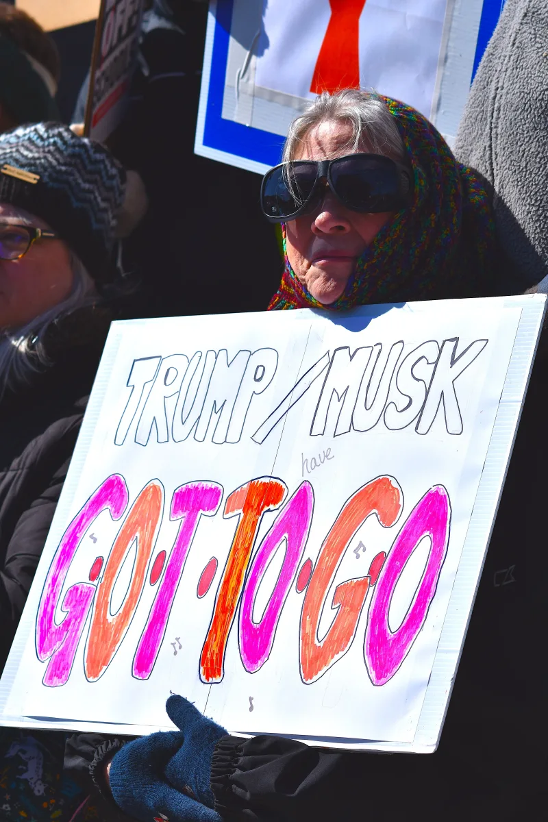 An older woman holds a protest sign against Trump and Musk outdoors in winter.