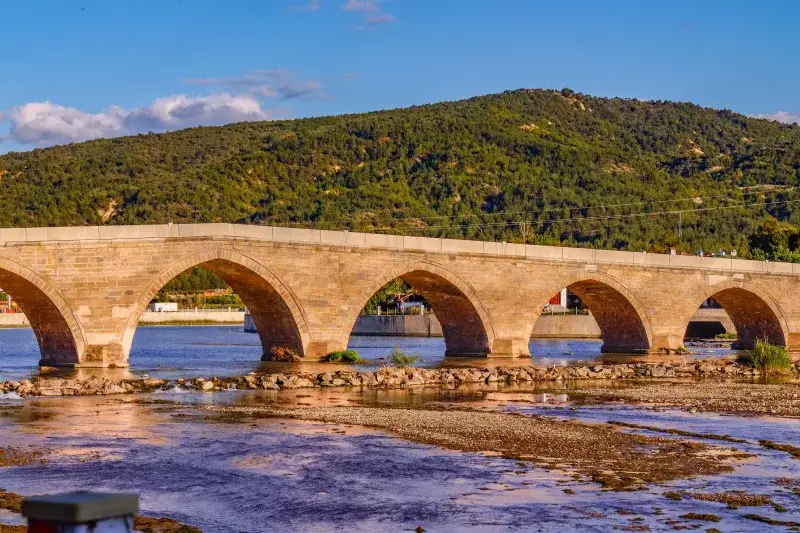 Stunning view of a historic stone arch bridge in Çorum, Türkiye surrounded by lush hills and calm river.