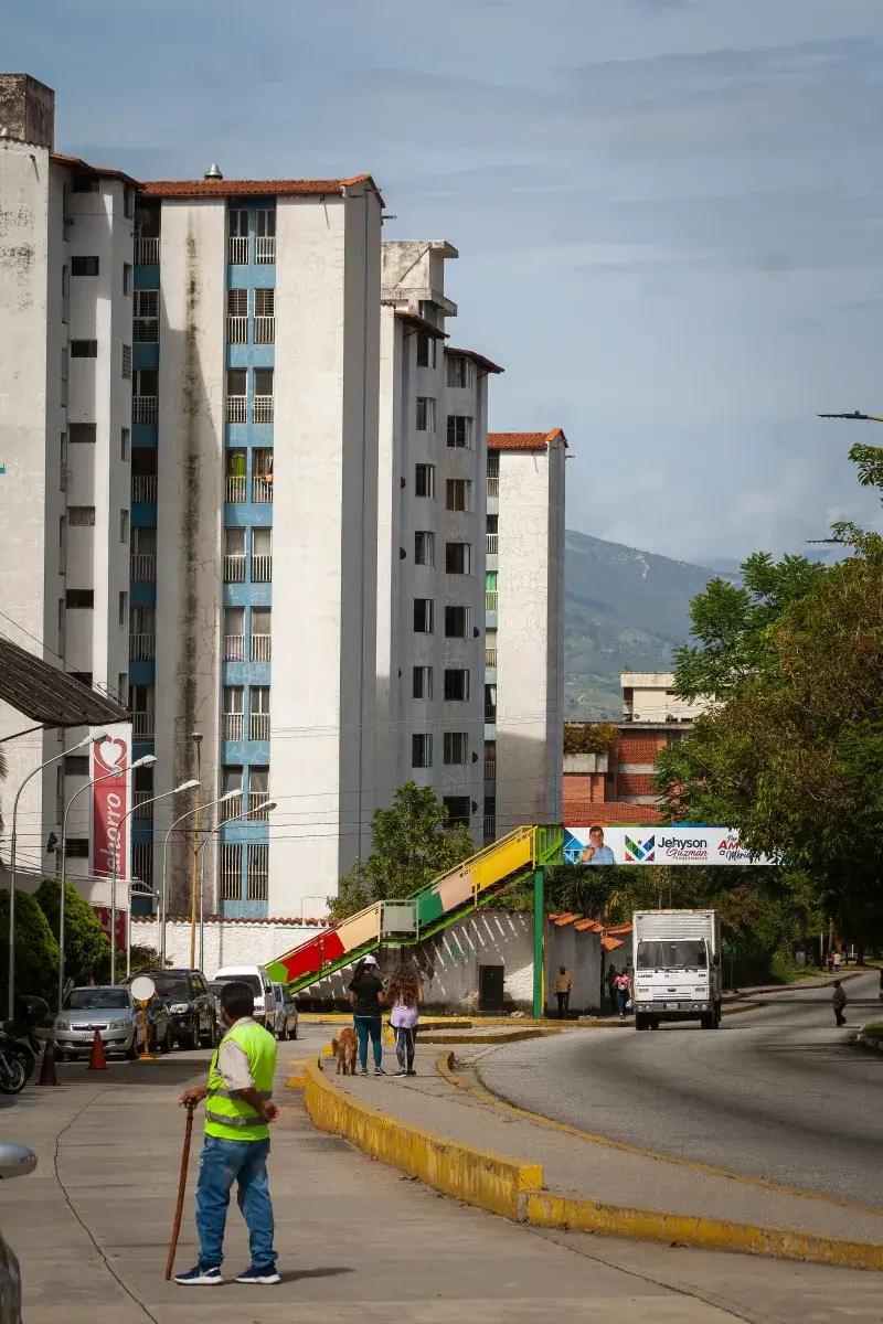 A lively urban scene in Venezuela featuring a colorful footbridge, high-rise residential buildings, and bustling street life.