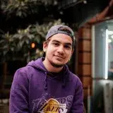 Casual portrait of a smiling young adult wearing a Lakers hoodie, sitting outdoors in urban Ankara.