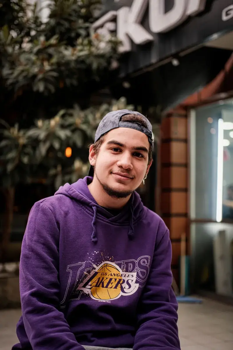 Casual portrait of a smiling young adult wearing a Lakers hoodie, sitting outdoors in urban Ankara.