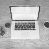 Monochrome image of a laptop, camera, lens, and coffee cup on a wooden desk