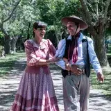 Smiling couple wearing traditional gaucho clothing walking in a lush park in Chascomús, Argentina.