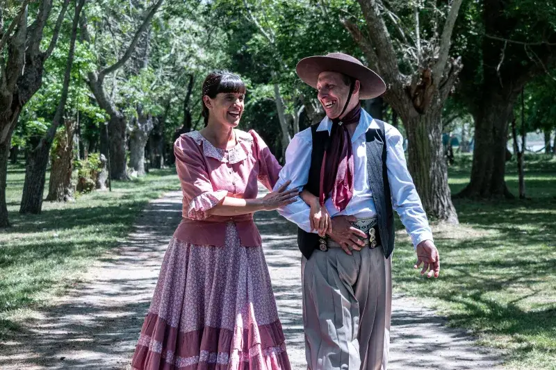 Smiling couple wearing traditional gaucho clothing walking in a lush park in Chascomús, Argentina.