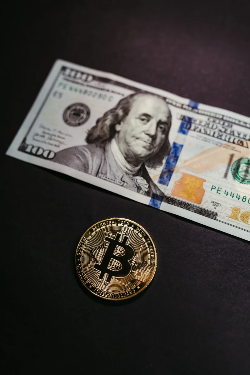 Close-up of a Bitcoin coin with a US 100 dollar bill, symbolizing digital and traditional currency.