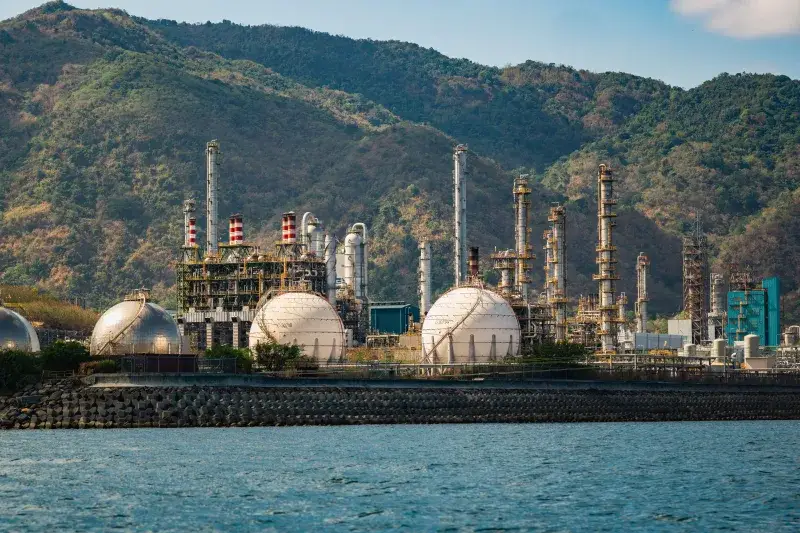 Refinería industrial con tanques esféricos junto a la costa, con un fondo de montañas.
