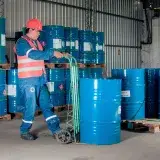 Industrial worker with a hand truck moving blue barrels in a warehouse setting.