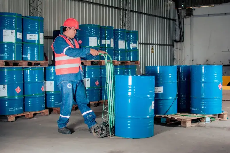 Trabajador industrial con un montacargas moviendo barriles azules en un almacén.