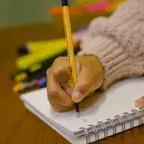 A person's hand writing in a spiral notebook using a pen, surrounded by colorful pens.