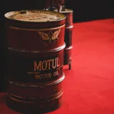 A close-up of a rusty oil barrel in an industrial setting with a red backdrop.
