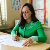 A female teacher checks papers at her desk in a classroom setting.