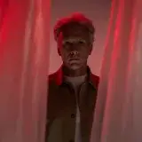 Elderly man with short gray hair looks through curtains under dramatic red lighting. Studio portrait.