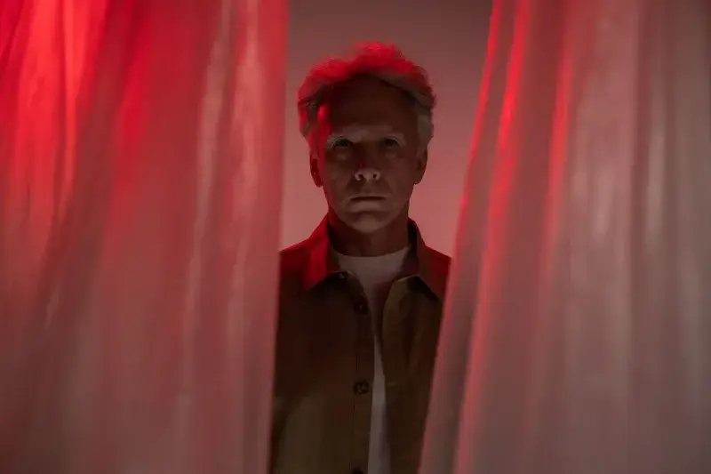 Elderly man with short gray hair looks through curtains under dramatic red lighting. Studio portrait.