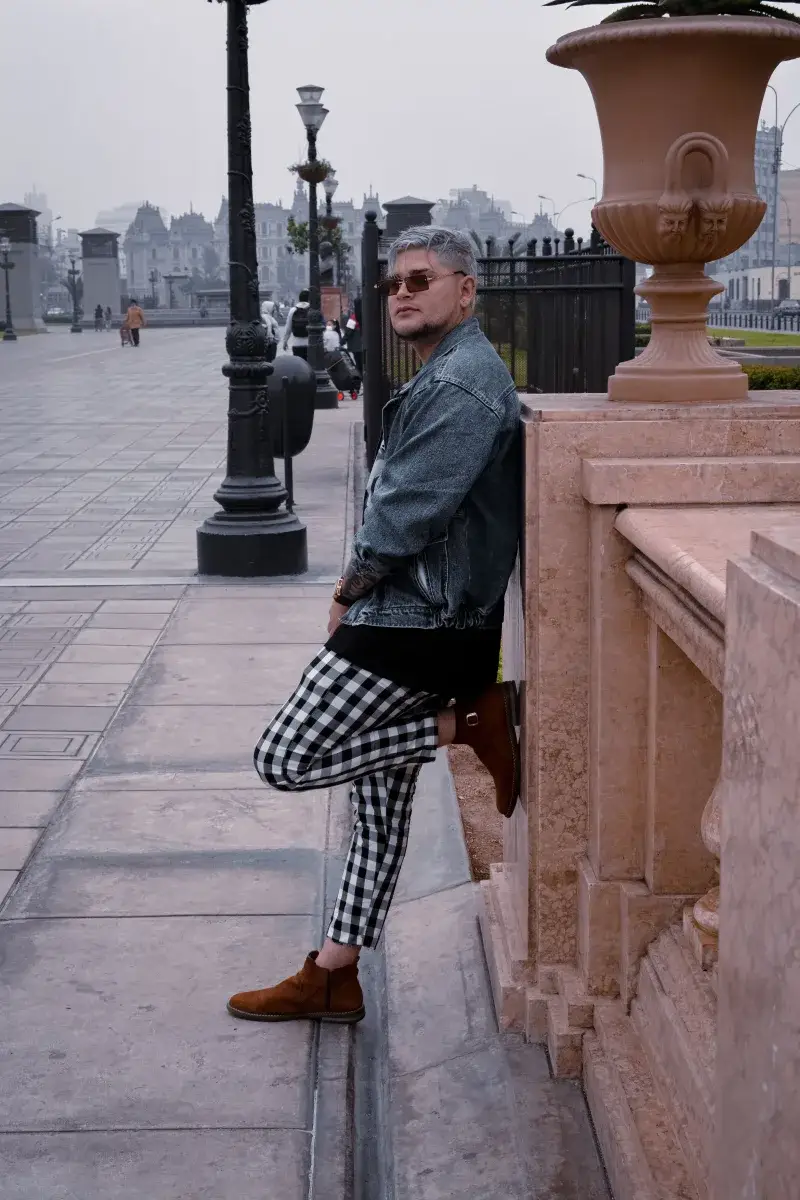 Man in denim jacket and checkered pants poses in urban Maracaibo, showcasing stylish fashion.