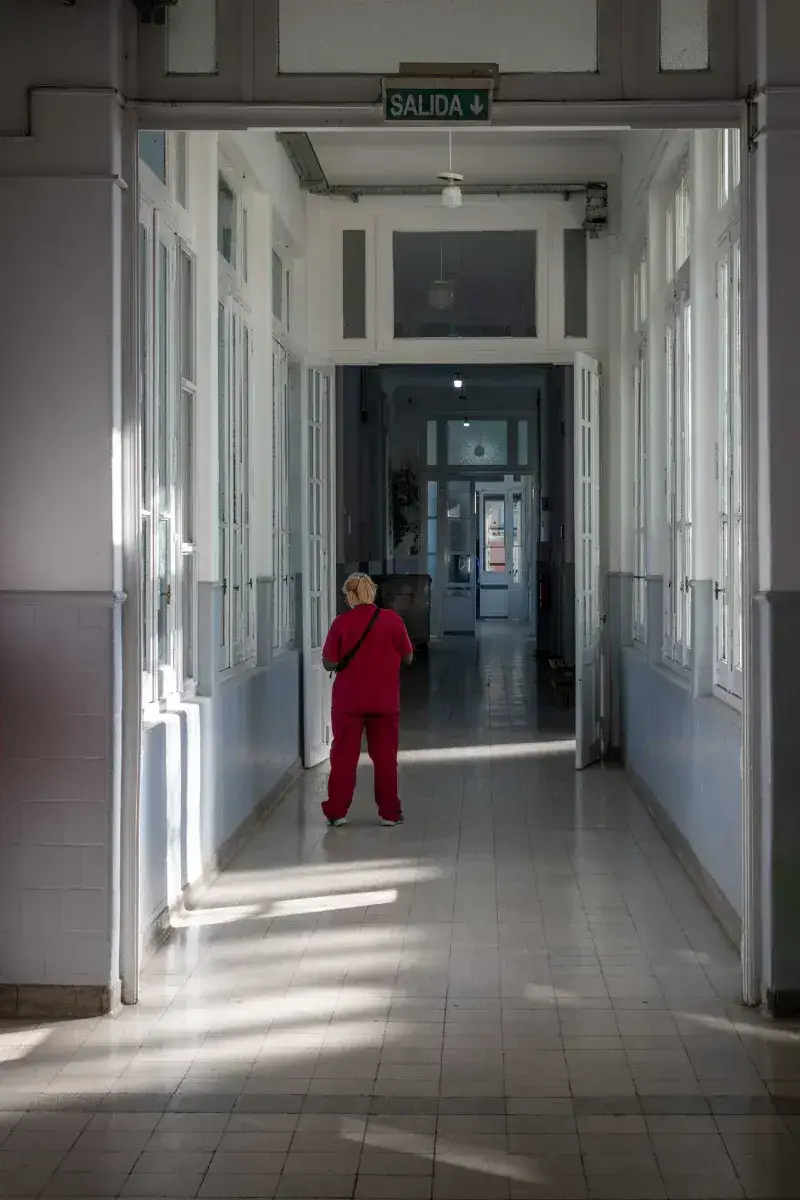 A nurse in red scrubs walks through a sunlit corridor in a hospital setting.