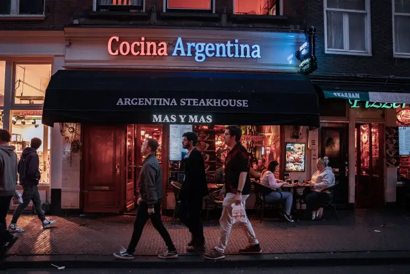 Evening view of pedestrians passing by a bustling Argentina Steakhouse in a lively urban setting.