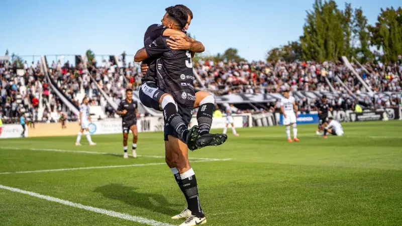 Two soccer players joyfully celebrate a goal during a match on a sunny day.