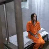 A woman sits in a prison cell wearing an orange jumpsuit with tattoos on her arms.