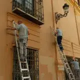 Three men on ladders working on the facade of a historic building in Spain, showcasing restoration efforts.