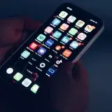 Close-up of hands holding a smartphone displaying various app icons in a dimly lit room.