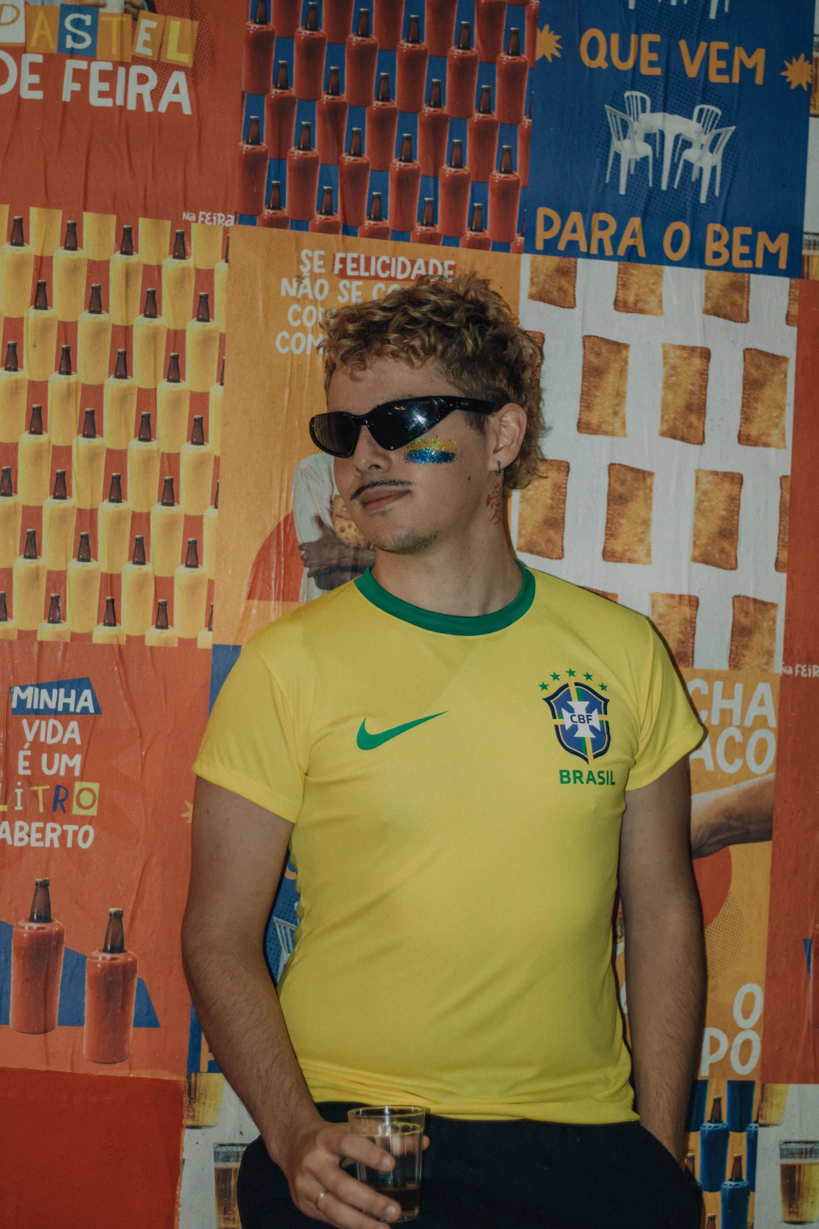 Joven fan brasileño de fútbol con la cara pintada se encuentra frente a un fondo de carteles coloridos sosteniendo una bebida.