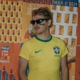 Joven fan brasileño de fútbol con la cara pintada se encuentra frente a un fondo de carteles coloridos sosteniendo una bebida.