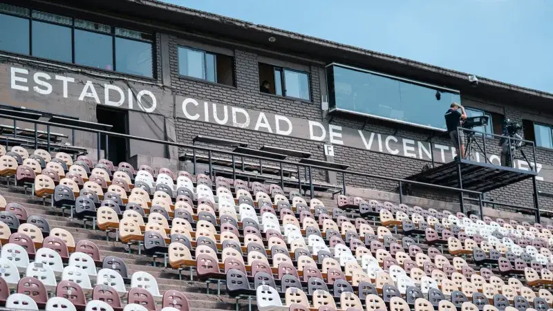 Empty stadium seats with a view of Estadio Ciudad de Vicente López sign, featuring filming equipment and operators.