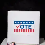 A ballot box with 'VOTE' sign, surrounded by voting stickers and a pen on a table.