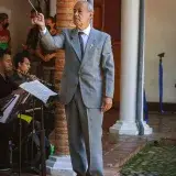 Senior conductor in gray suit leading an orchestra outdoors in Venezuela.