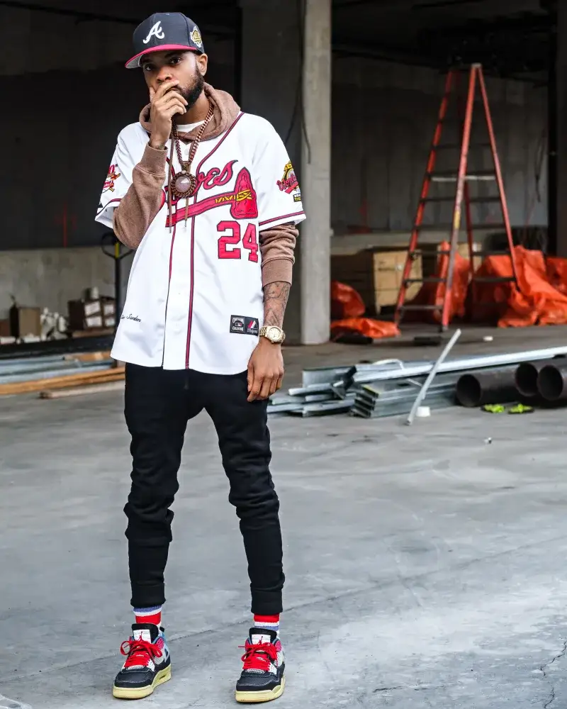 African American man posing stylishly in Atlanta Braves jersey at a construction site. Urban fashion captured outdoors.