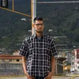 A man wearing a checkered shirt stands confidently outdoors in Venezuela with a scenic mountain backdrop.