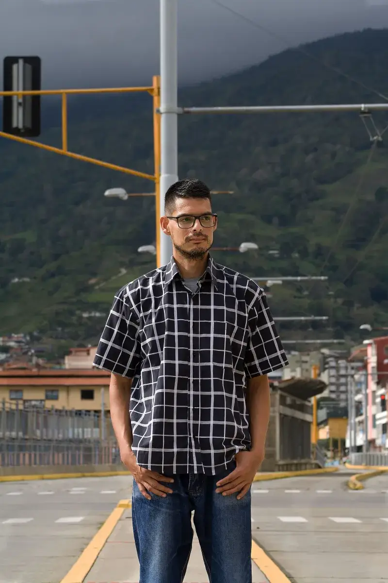 A man wearing a checkered shirt stands confidently outdoors in Venezuela with a scenic mountain backdrop.