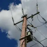 Low-angle view of a utility pole and power lines set against a blue sky with white clouds.