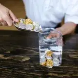 Hand placing gold and silver coins into a glass jar on a wooden table indoors.