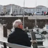 An elderly man sits peacefully overlooking a harbor in Nuova Aquitania, Francia, on a calm day.