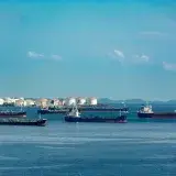 A fleet of cargo ships docked near oil storage tanks along a serene coastline with a clear blue sky above.