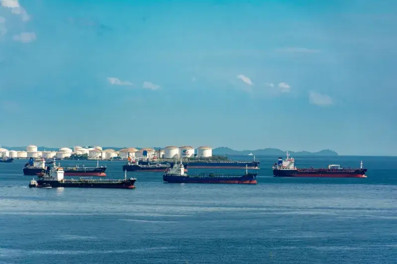 A fleet of cargo ships docked near oil storage tanks along a serene coastline with a clear blue sky above.