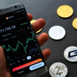 Close-up of a smartphone app showing Bitcoin trading details with crypto coins on a black surface.