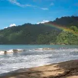 Vibrant rainbow stretches over a lush green hill by a sandy beach in Trinidad and Tobago.