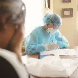 Medical professionals in PPE working at a desk during a daytime setting indoors.