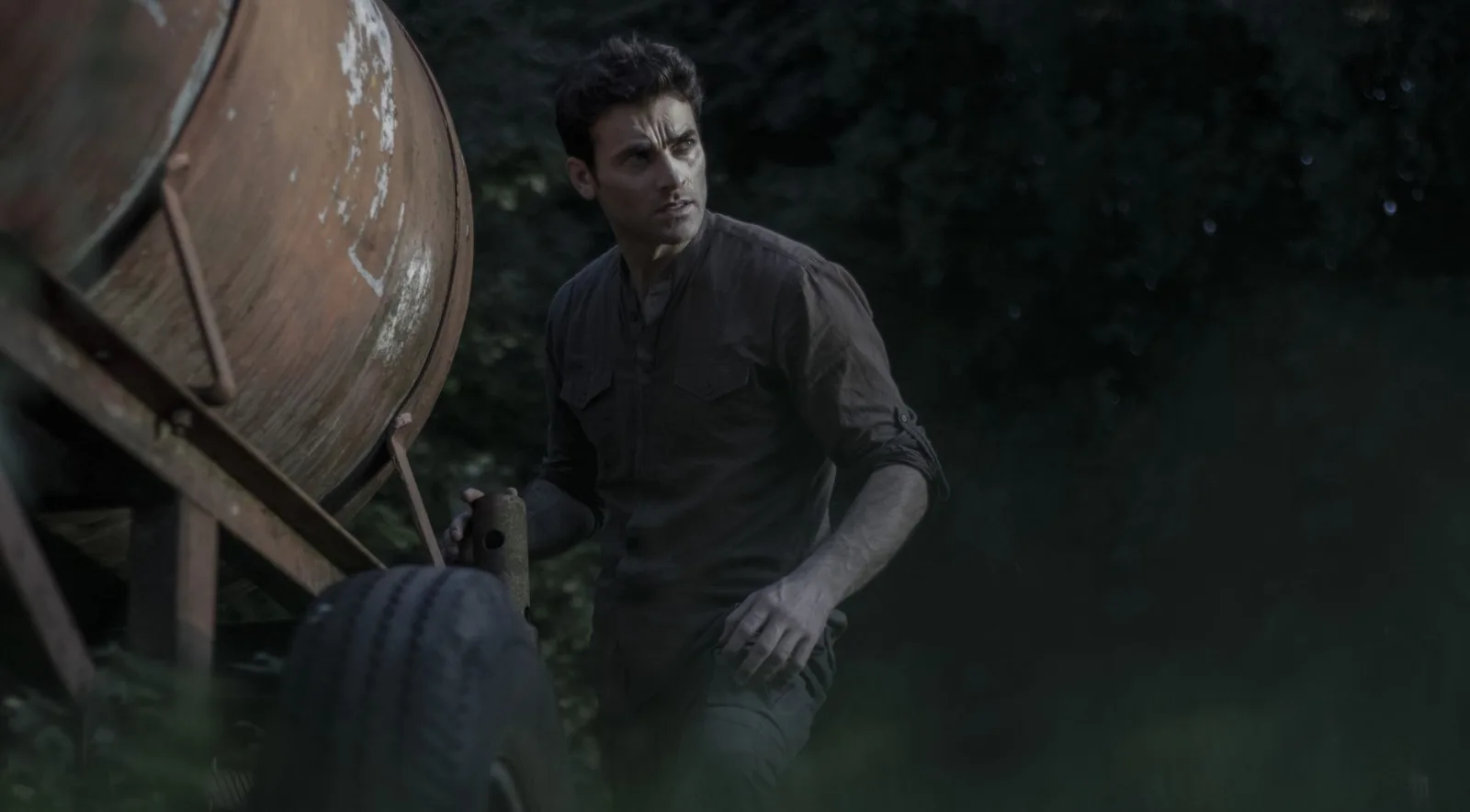 A man with short hair stands beside a rustic trailer in a forest setting.