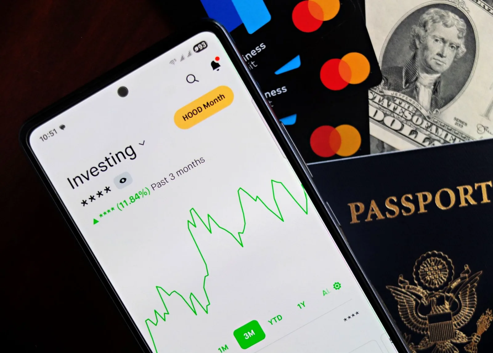 Smartphone displaying investing app, with credit cards, cash, and passport nearby, symbolizing finance