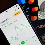 Smartphone displaying investing app, with credit cards, cash, and passport nearby, symbolizing finance