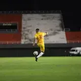 Soccer player in yellow jersey kicking a ball on a green pitch during night match under lights.