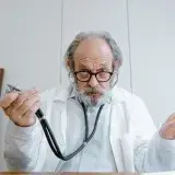 Senior doctor gesturing with stethoscope in a clinical setting, emphasizing telemedicine.